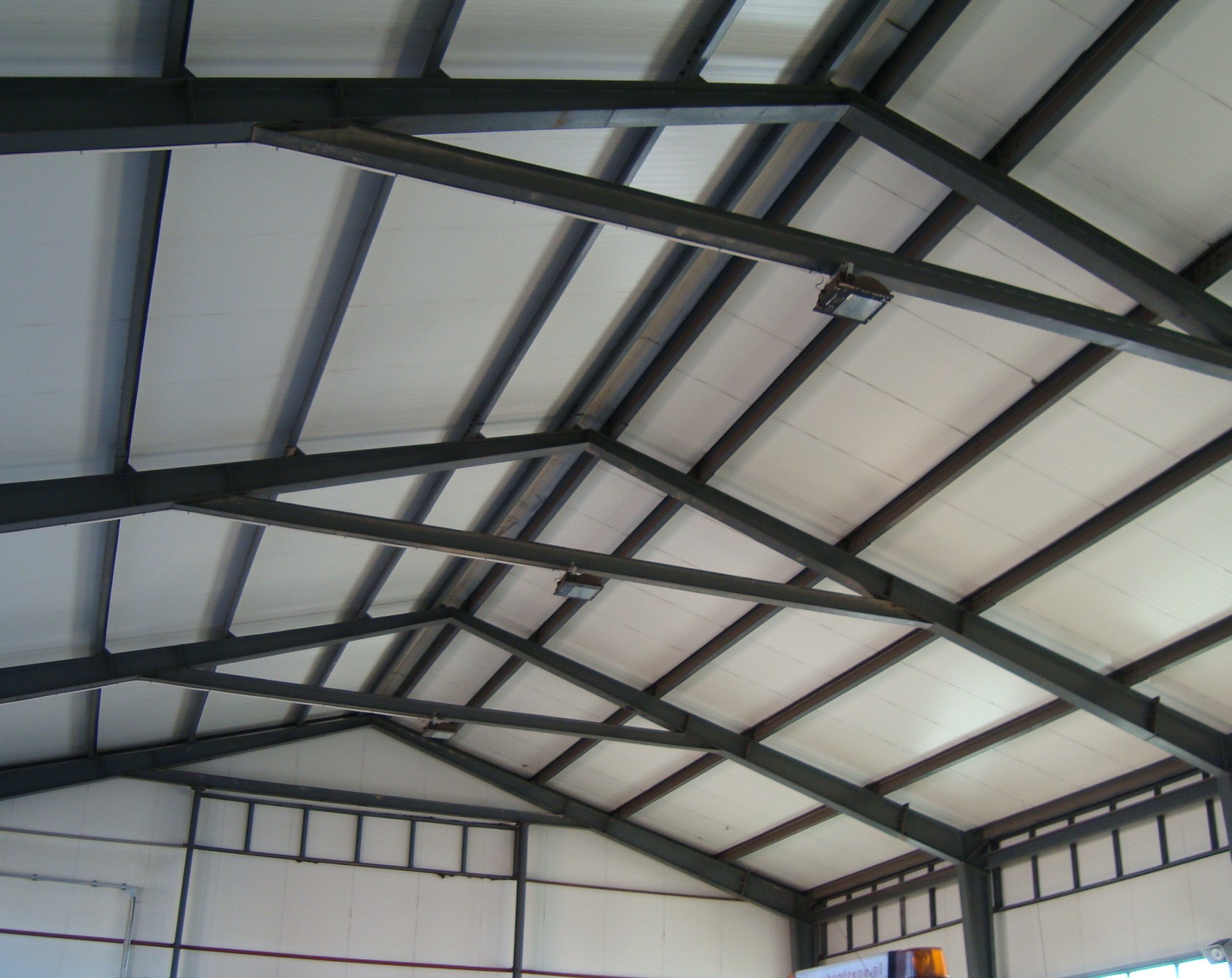 Wnętrze hali produkcyjnej z widoczną konstrukcją stalową dachu i oświetleniem punktowym, widok od dołu.